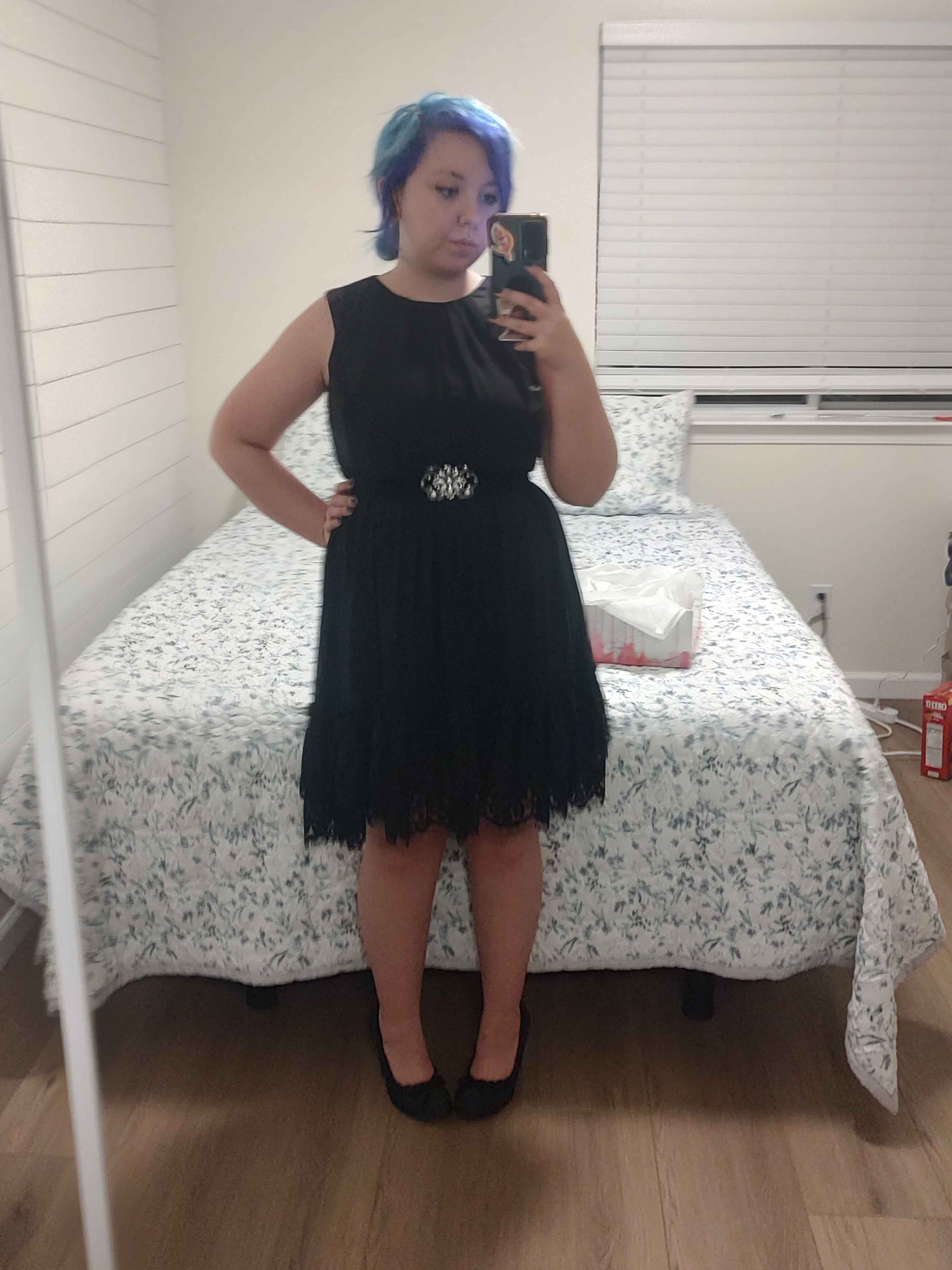 A person with purple and blue hair stands in front of a mirrored closet door. They are wearing a black, knee-length, sleeveless dress and black high heels. The dress has lace at the bottom of a sheer skirt laying over a much tighter inner slip, and the bodice has two different textures of fabric. The center fabric is a matte texture, and the outer fabric has a satin look. The dress has a velvet-look belt with an ornate silver ornament at the center. In the background is a queen-sized bed with a white and purple floral comforter. On the bed is an open shoebox. 